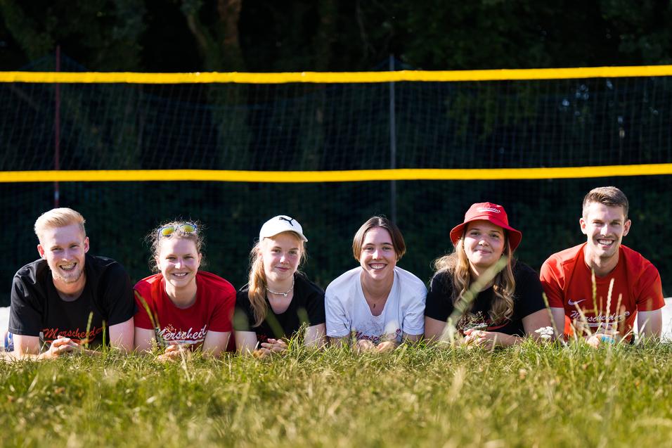 Sechs Personen liegen im Gras vor einem Beachvolleyballfeld mit gelben Netzen und Bäumen im Hintergrund.